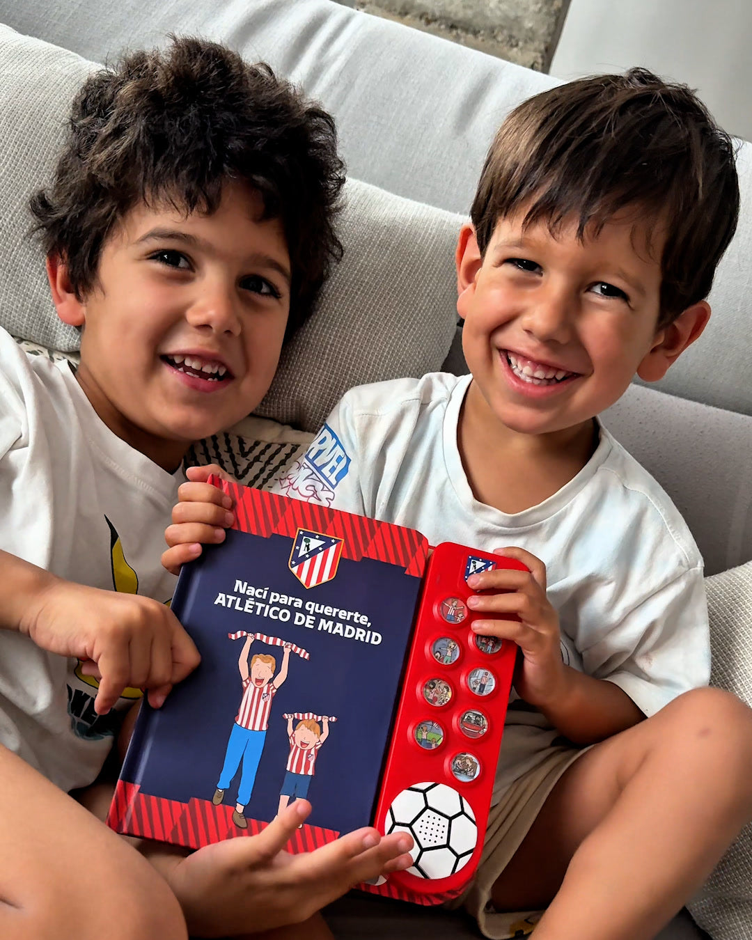 Two boys on a sofa holding up the Atletico de Madrid Sound Book, both smiling, the crest and the eight sound buttons in view.
