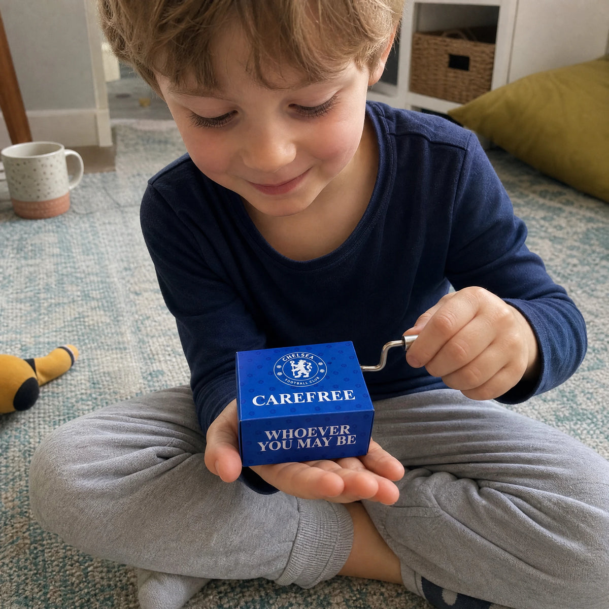 Un bambino seduto a gambe incrociate su un tappeto tiene il carillon del Chelsea FC sul palmo aperto di una mano e gira la piccola manovella con l'altra, sorridendo mentre suona.