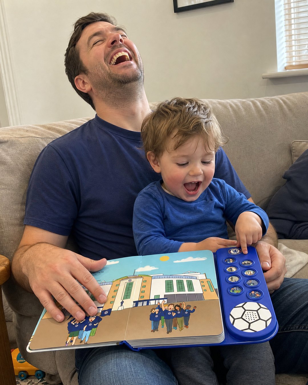 A dad on the sofa with his toddler on his lap, the boy pressing a sound button on the open Chelsea Sound Book.