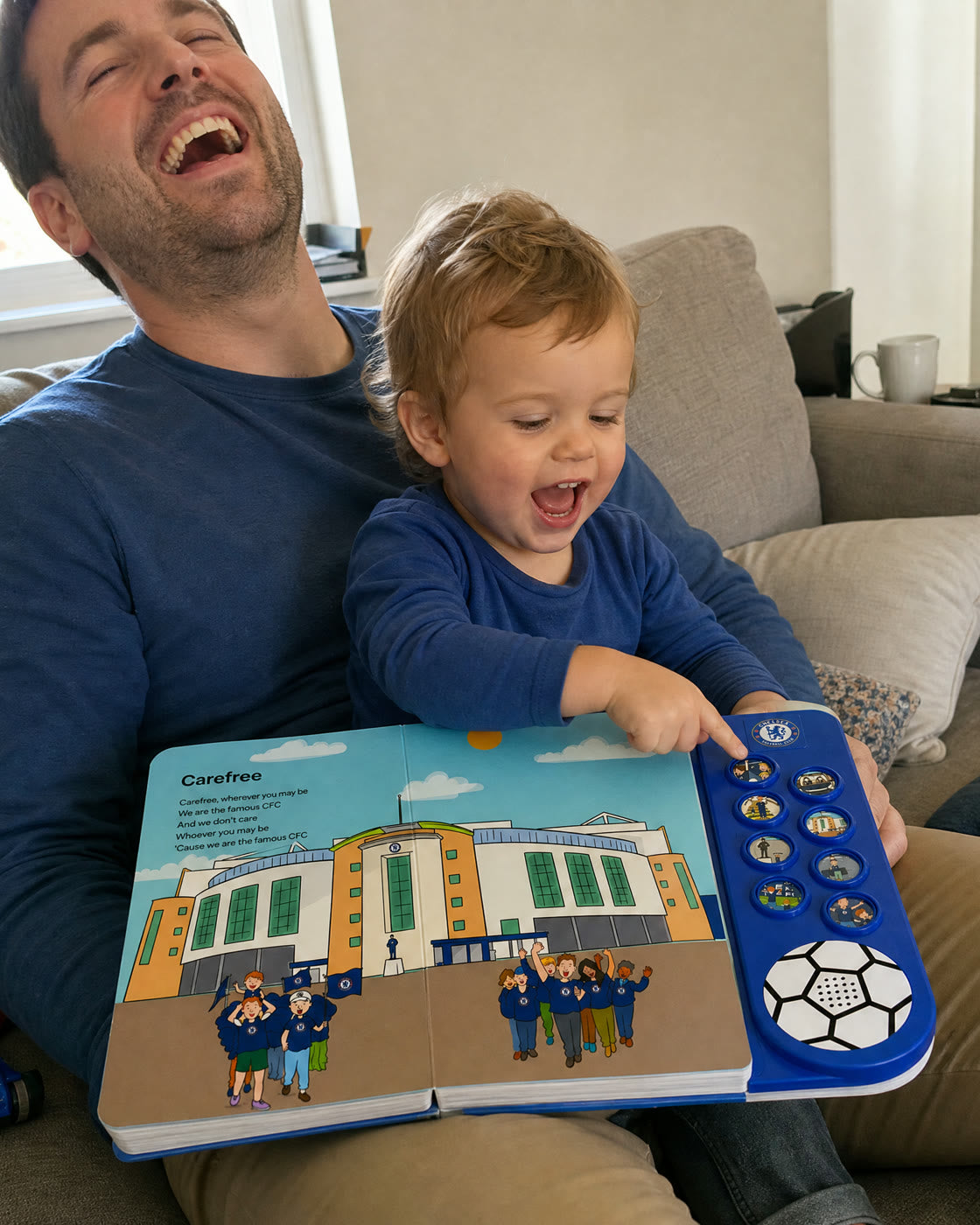 A laughing dad with a toddler on his lap pressing a button on the Chelsea Sound Book