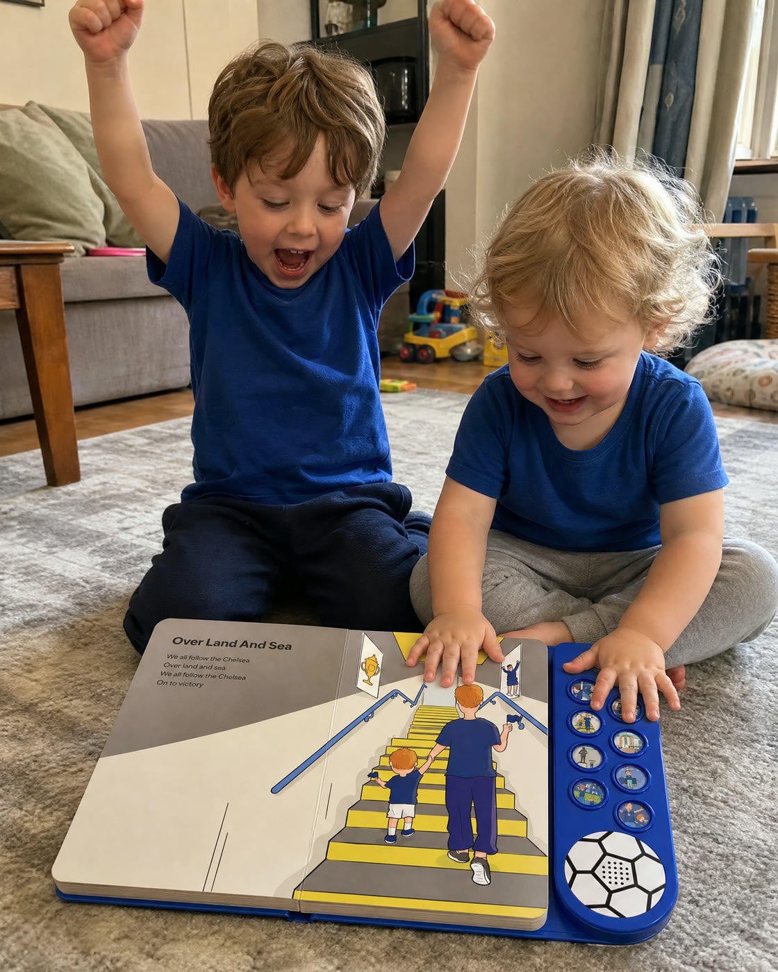Two young siblings on a living-room rug with the Chelsea Sound Book open on the Over Land And Sea page, the older one cheering with both fists up