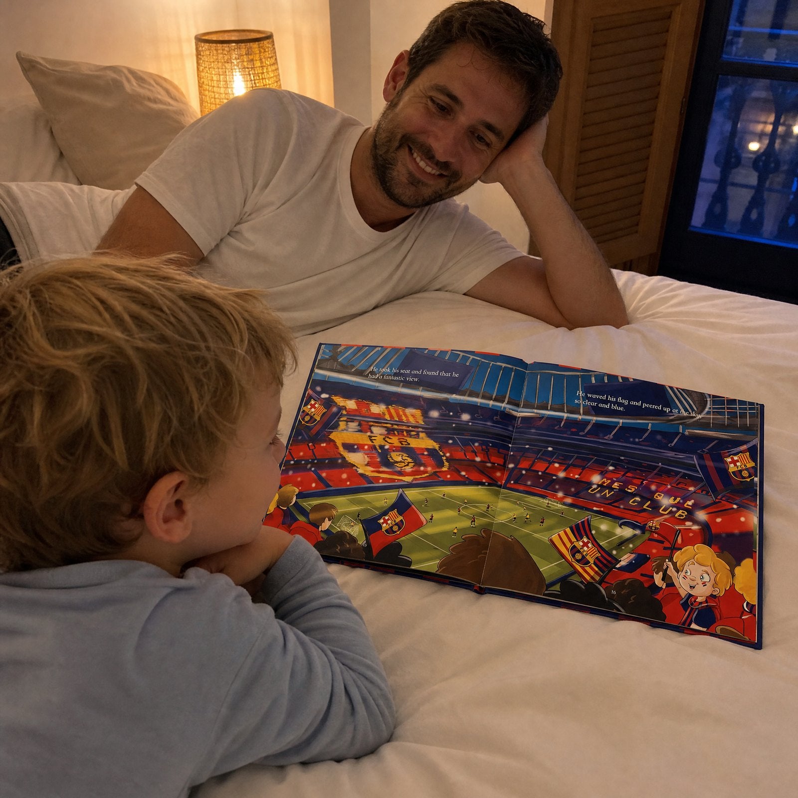 A young boy lying on his bed at night reading the FC Barcelona Personalised Book while his dad watches him and smiles.