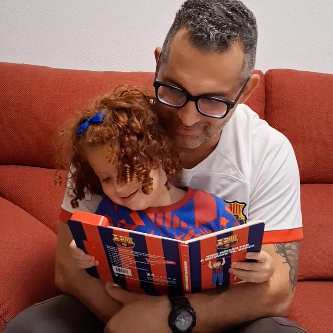 A dad with his daughter on his lap, both looking down at the open FC Barcelona Sound Book.