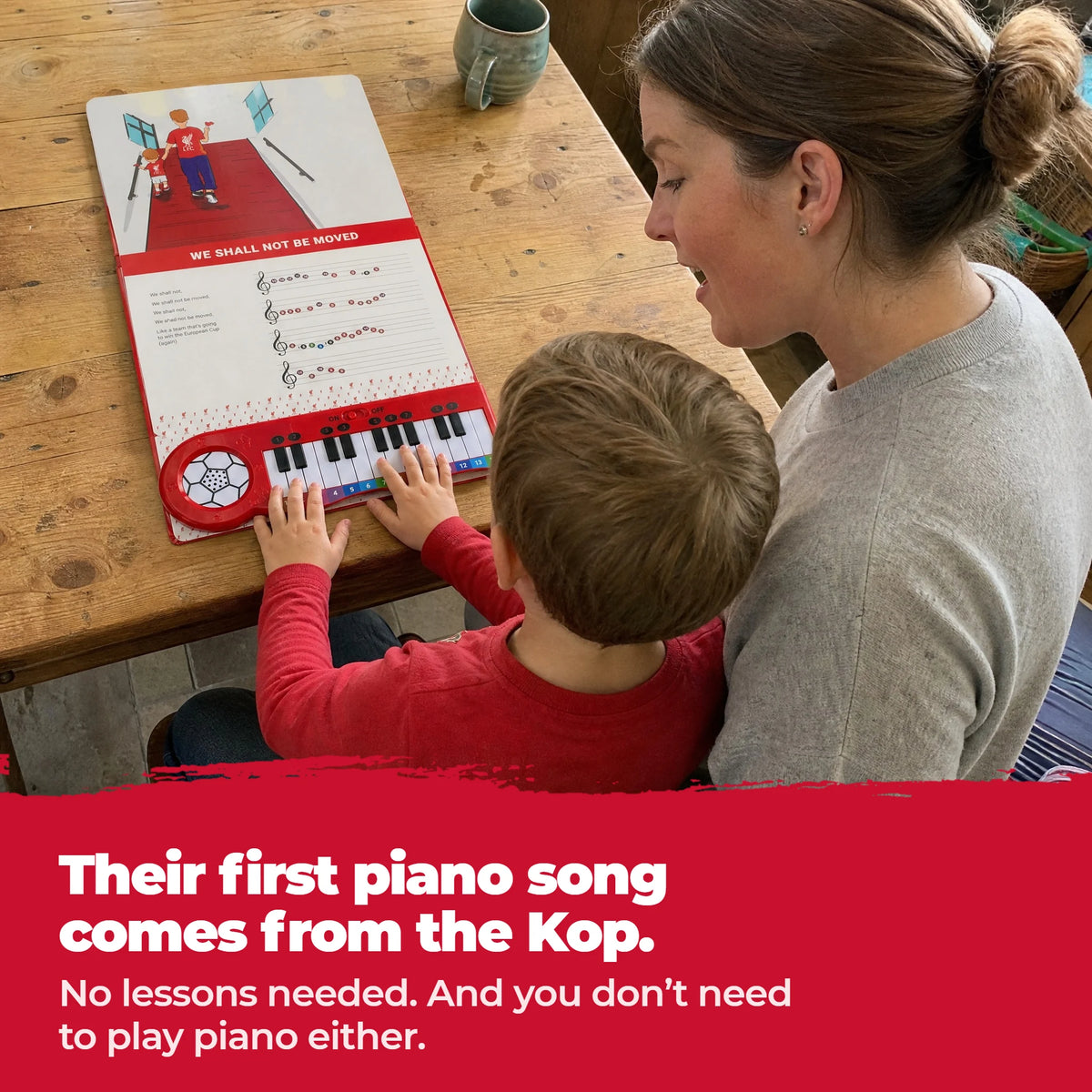 Un bambino in braccio alla mamma, al tavolo, suona i tasti del Libro di Pianoforte del Liverpool, aperto e steso davanti a loro.