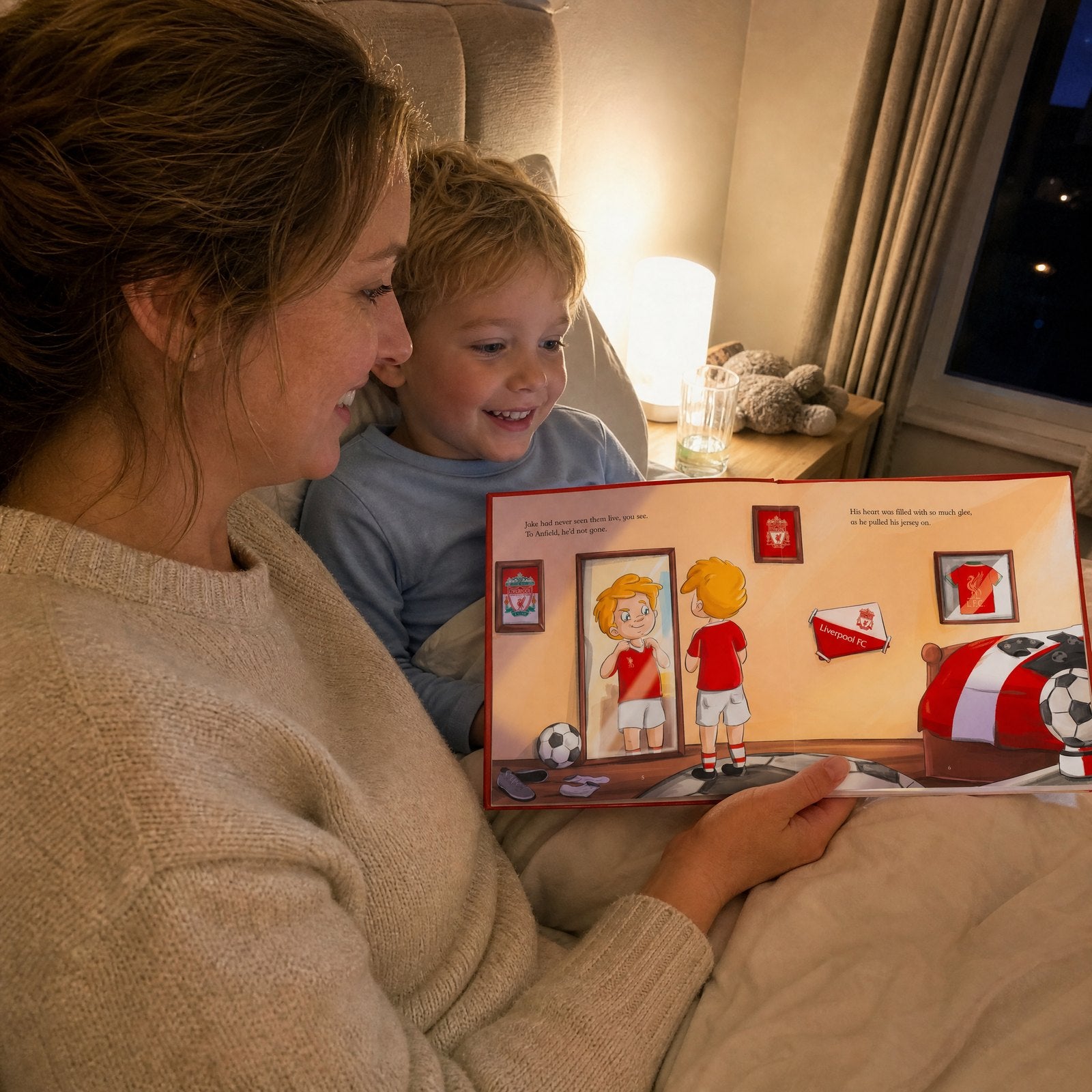 A mum and her young son in bed at night reading the Liverpool FC Personalised Book together, the boy grinning at the page.