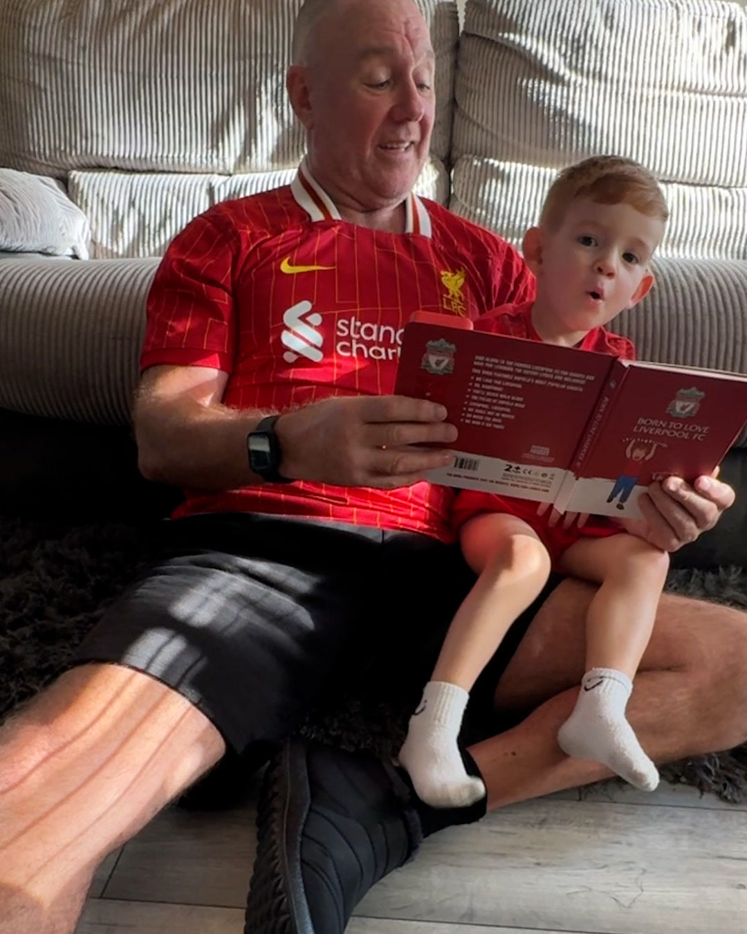 A grandad in a Liverpool shirt with his grandson on his lap, both singing along to the open Liverpool Sound Book.