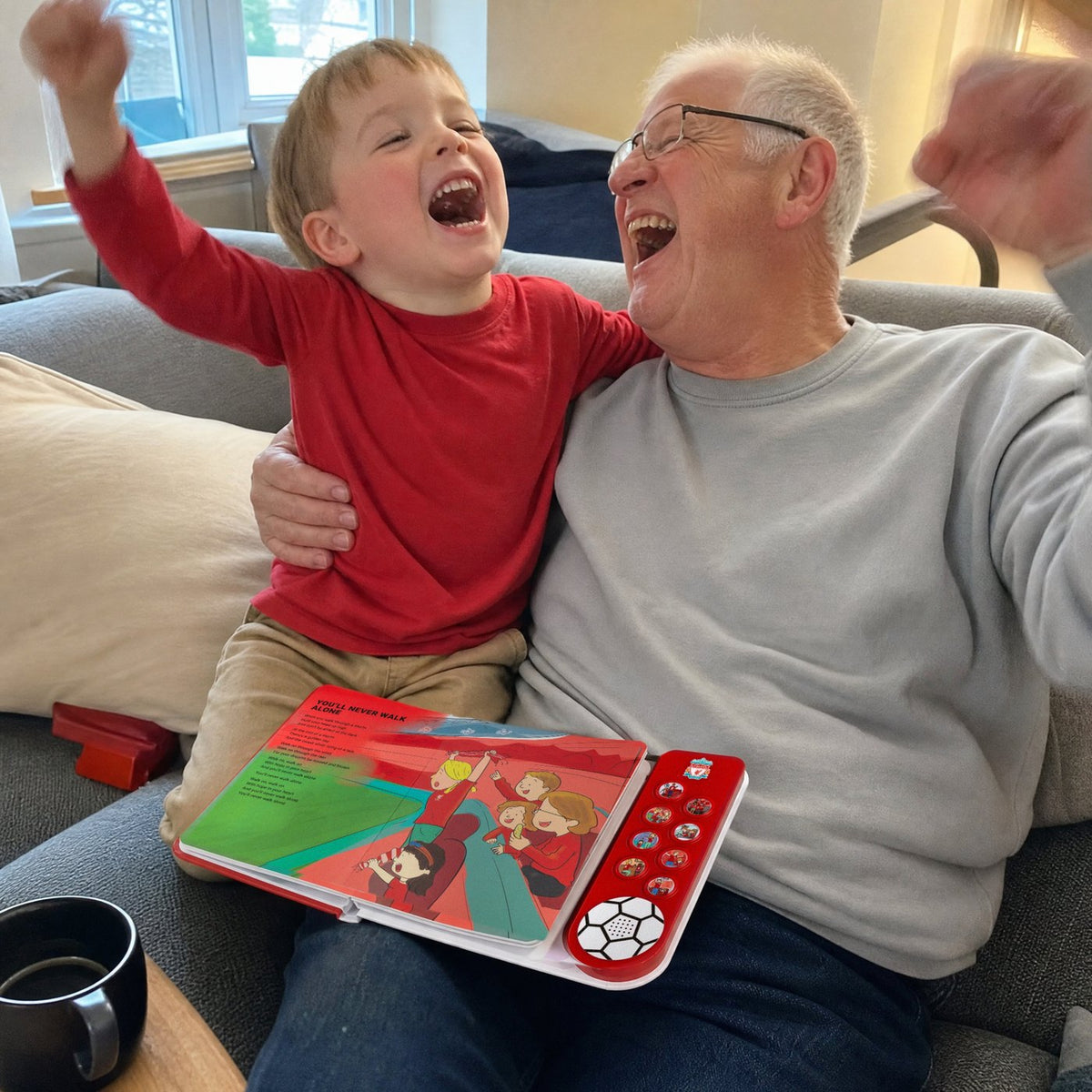 A grandad and his grandchild sing together on the sofa, the Liverpool FC Sound Book open on his knee