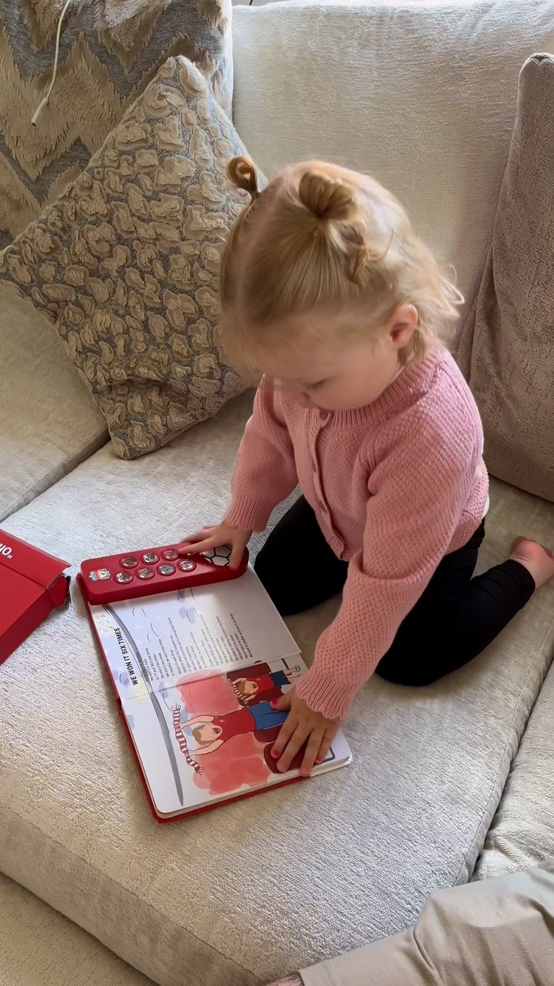 A toddler kneels on a sofa, pressing the buttons of the Liverpool FC Sound Book on her own