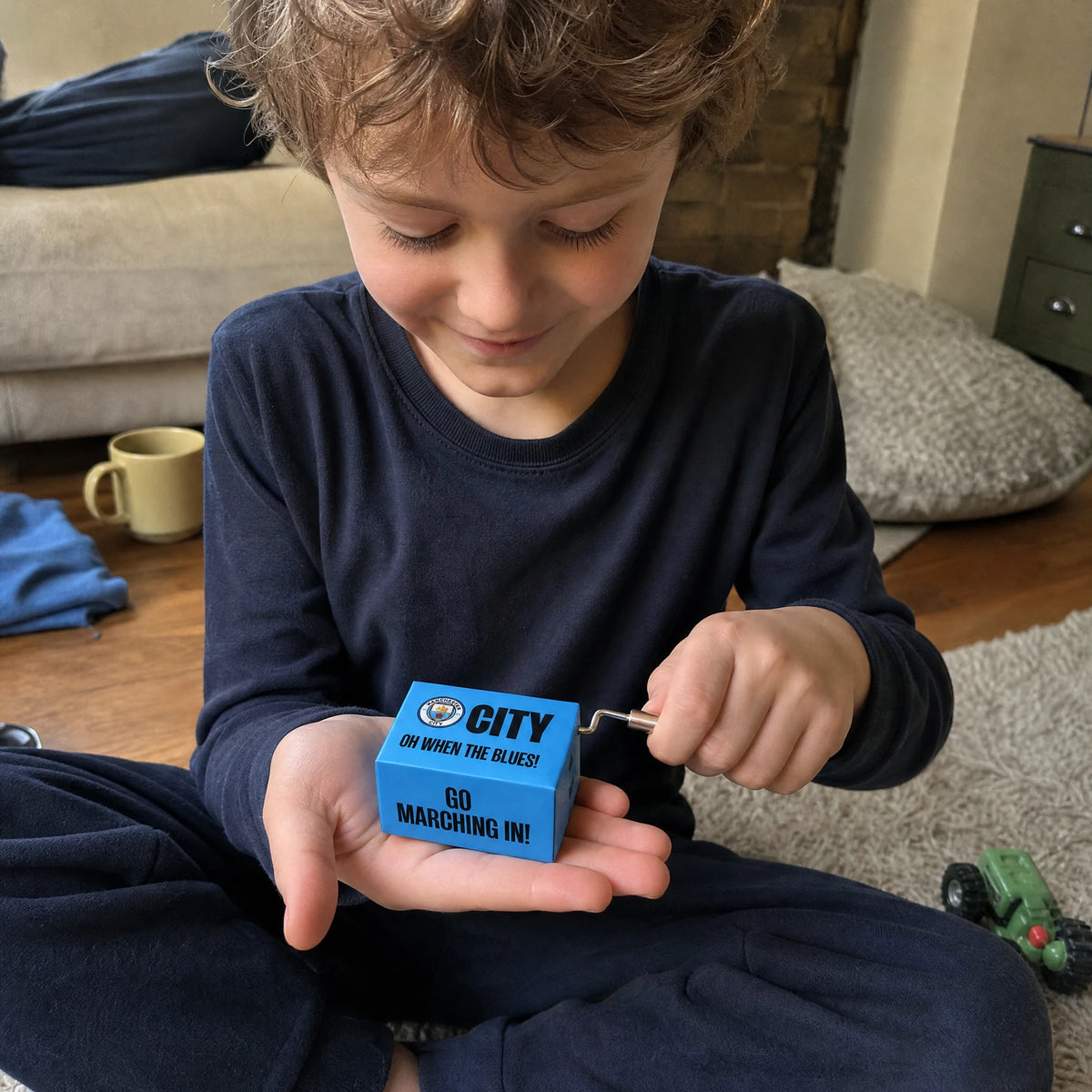 Un niño sentado con las piernas cruzadas sobre una alfombra sostiene la caja de música del Manchester City en la palma de una mano y gira la pequeña manivela con la otra, sonriendo mientras suena.