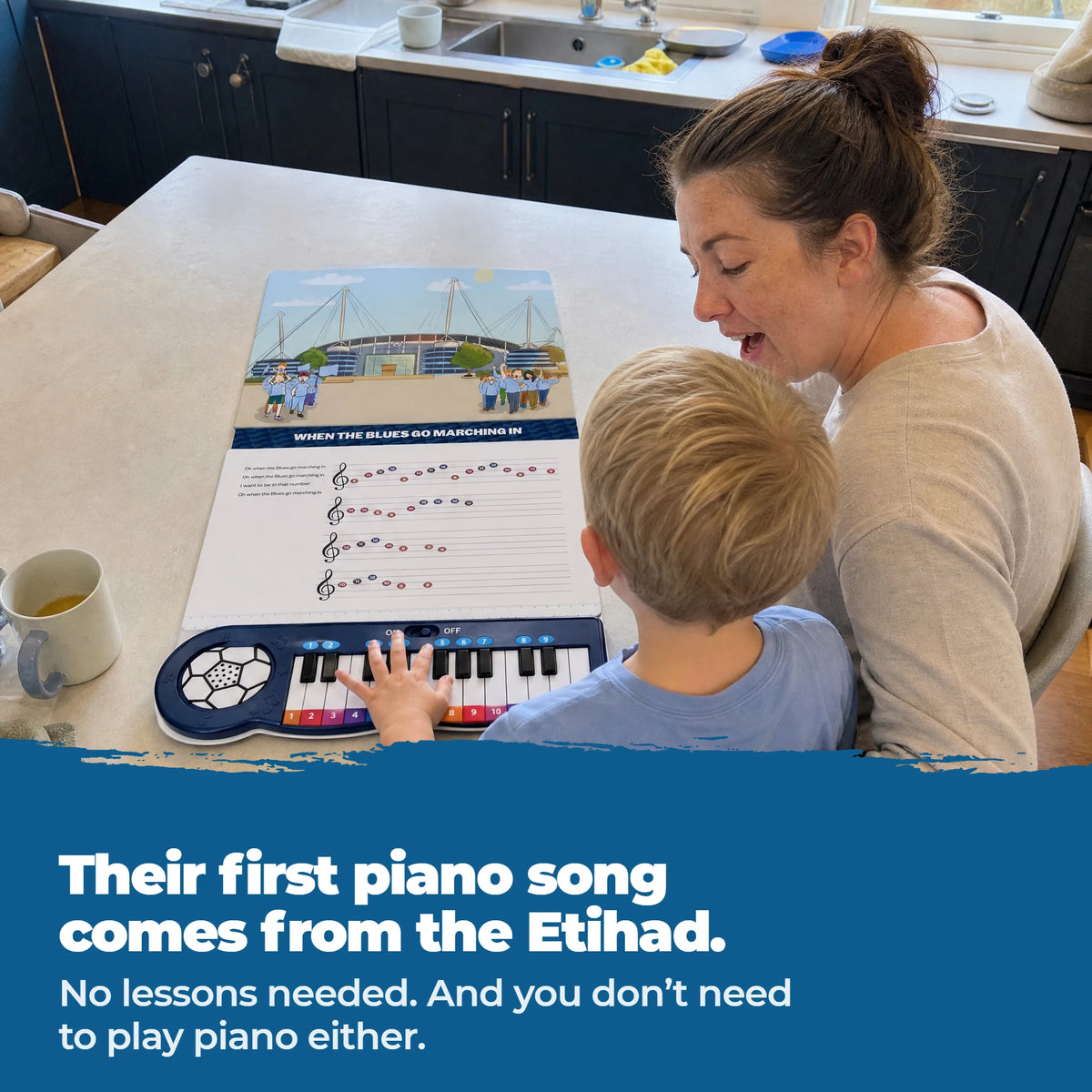 A young child on a parent's lap at a table, playing the keys of the Manchester City Piano Book, which lies open flat in front of them.