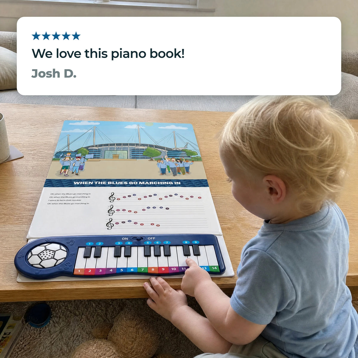 A toddler at a low table playing the keys of the Manchester City Piano Book, which lies open flat, beneath a five-star review from Josh D.: "We love this piano book!"