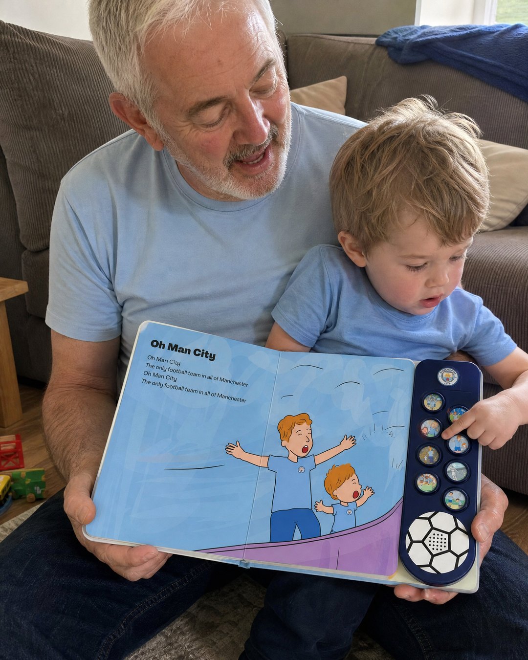 A grandad on the sofa with his grandson, the boy pressing a sound button on the open Man City Sound Book.
