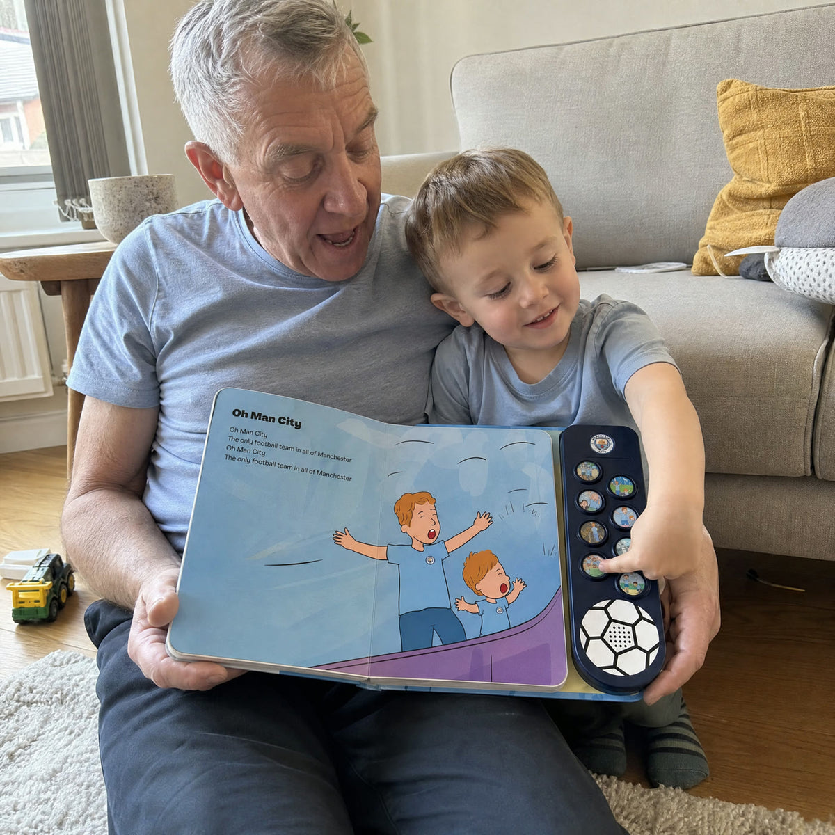 A grandfather in a pale sky-blue T-shirt on the living-room floor with a small boy, the Manchester City Sound Book open across their laps at Oh Man City, the boy's finger on a sound button.