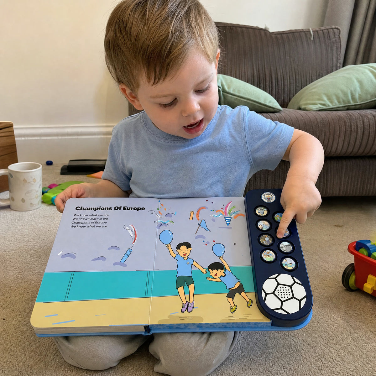 A toddler in a pale sky-blue top kneeling over the Manchester City Sound Book, open at Champions Of Europe, pressing one of the sound buttons.