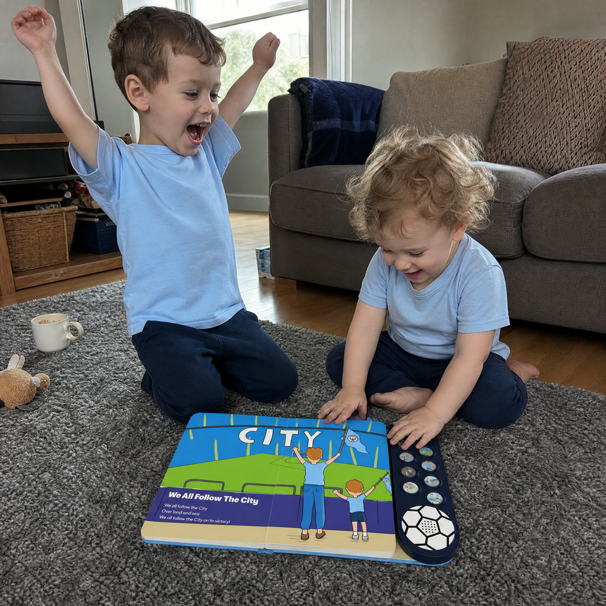 Two young siblings in pale sky-blue tops on a living-room rug: the older one kneeling with both fists in the air mid-chant, the younger one grinning down at the Manchester City Sound Book open at We All Follow The City.