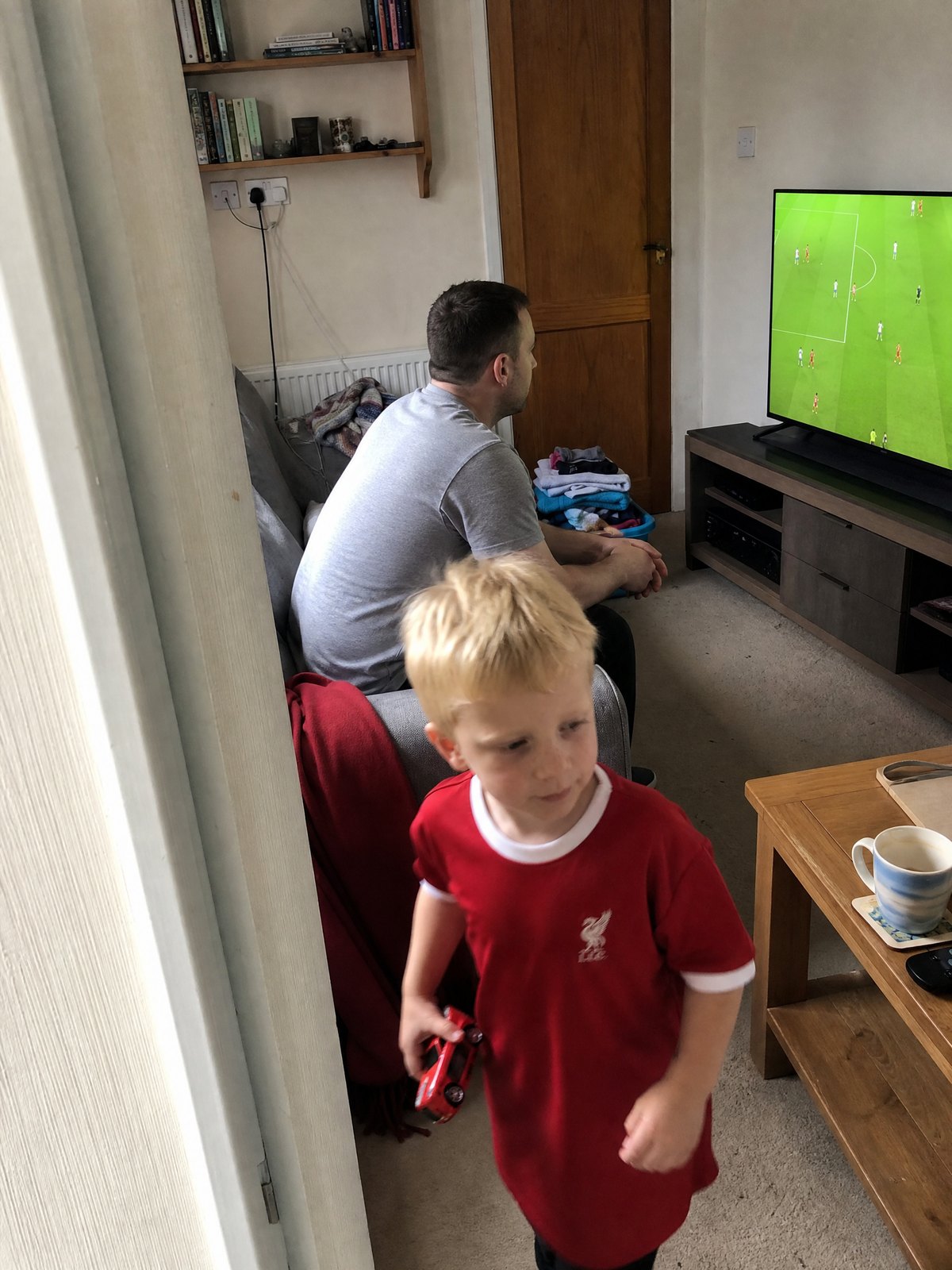 A dad on the edge of the sofa watching the match while his son wanders past