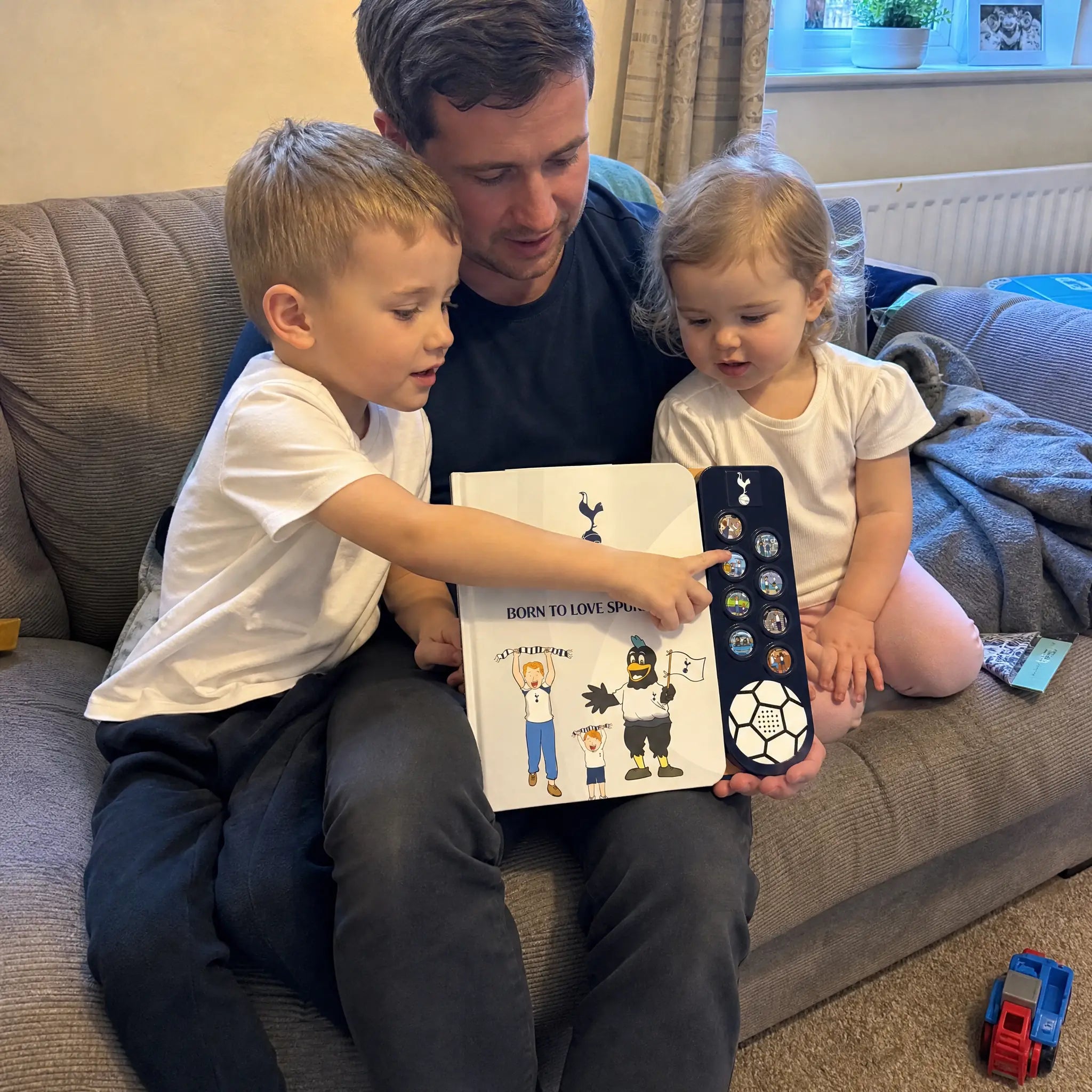A dad on the sofa with two children, one pointing at a sound button on the open Tottenham Hotspur Sound Book.
