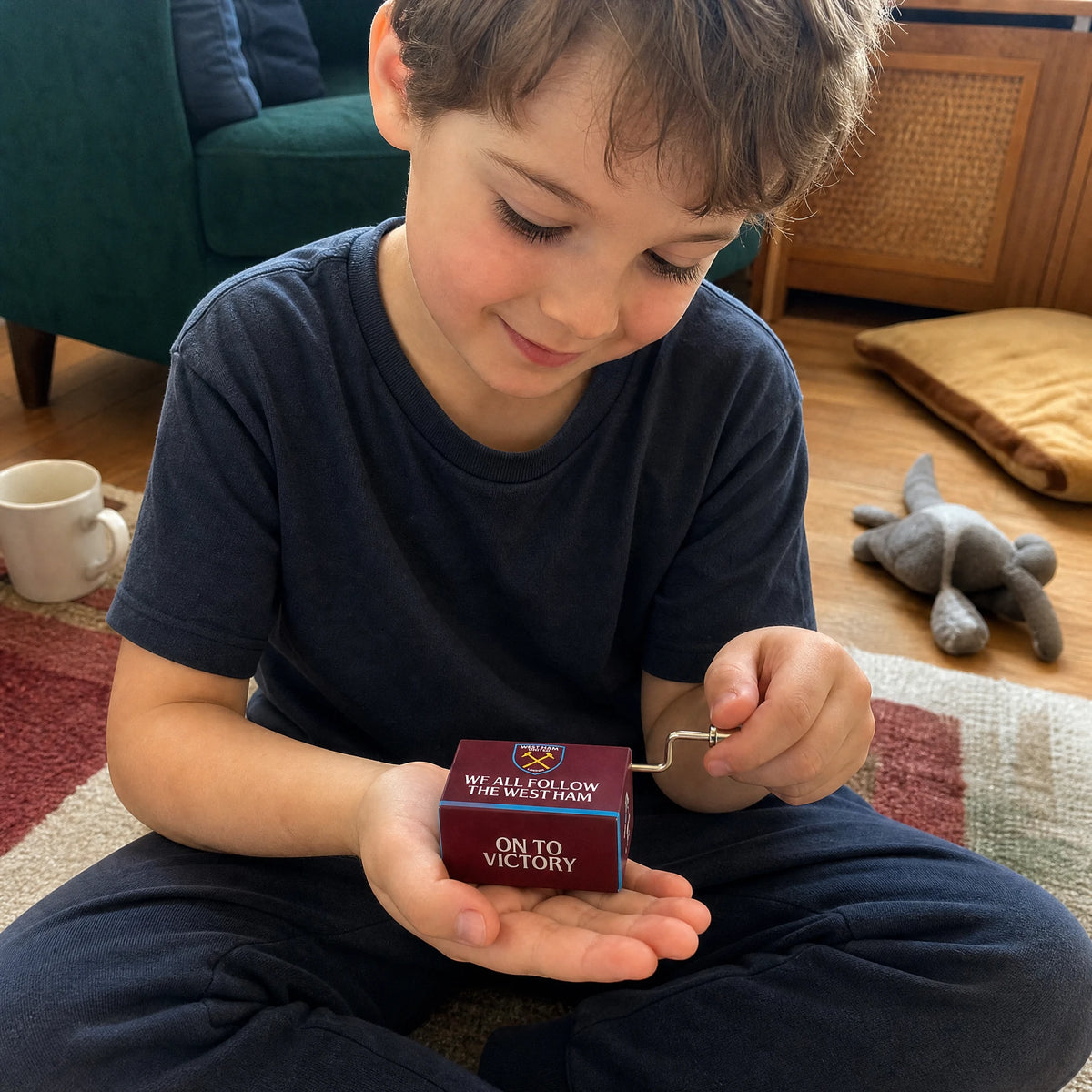 Ein Kind sitzt im Schneidersitz auf einem Teppich, hält die West-Ham-United-Spieluhr auf der offenen Handfläche und dreht mit der anderen Hand die kleine Kurbel, lächelnd, während sie spielt.