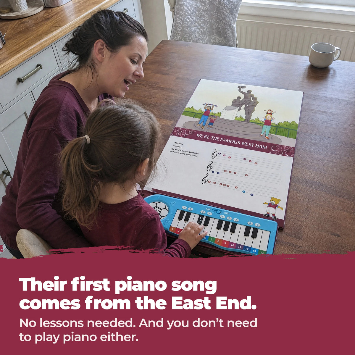 A young child on a parent's lap at a table, playing the keys of the West Ham United Piano Book, which lies open flat in front of them.