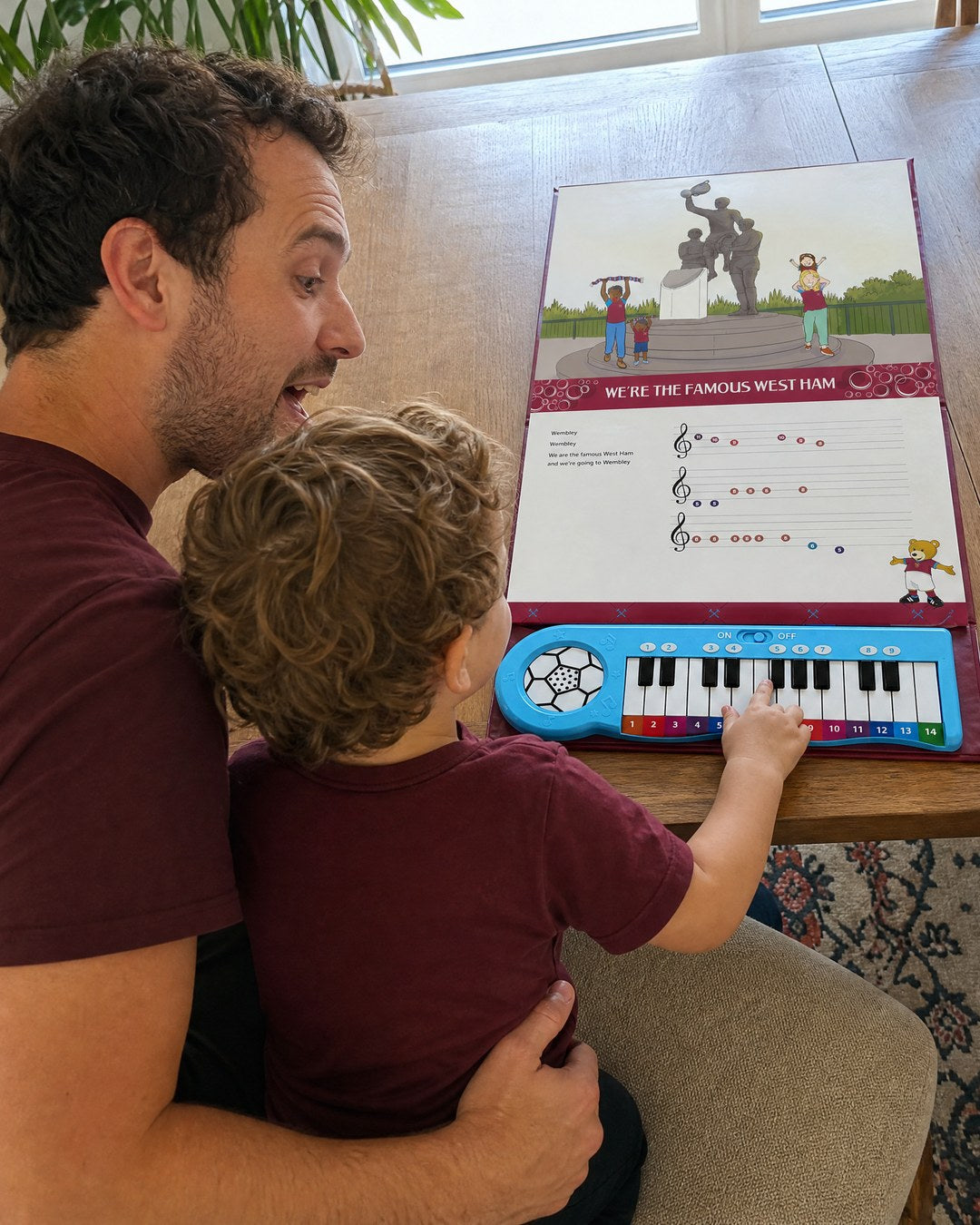 A dad at the dining table with his son on his lap, the boy playing the keys of the West Ham Piano Book lying open flat in front of them.