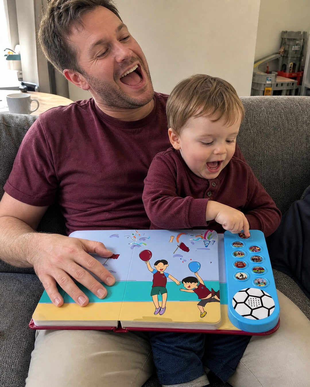A dad on the sofa with his toddler on his lap, the boy pressing a sound button on the open West Ham Sound Book.