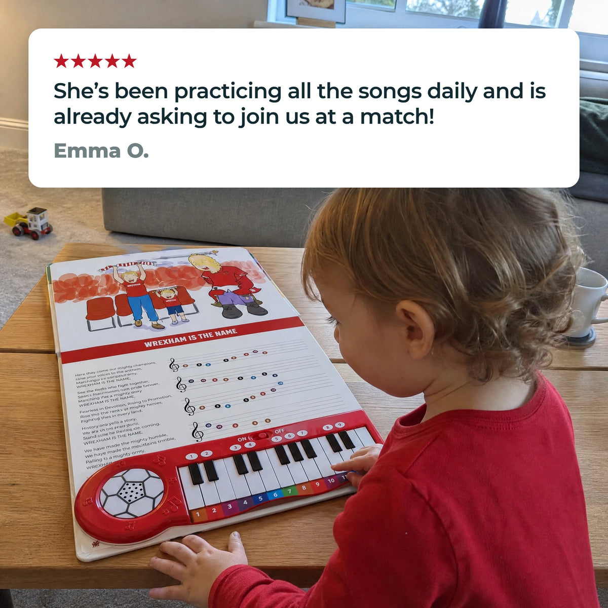 A toddler at a low table playing the keys of the Wrexham AFC Piano Book, which lies open flat, beneath a five-star review from Emma O.: "She’s been practicing all the songs daily and is already asking to join us at a match!"