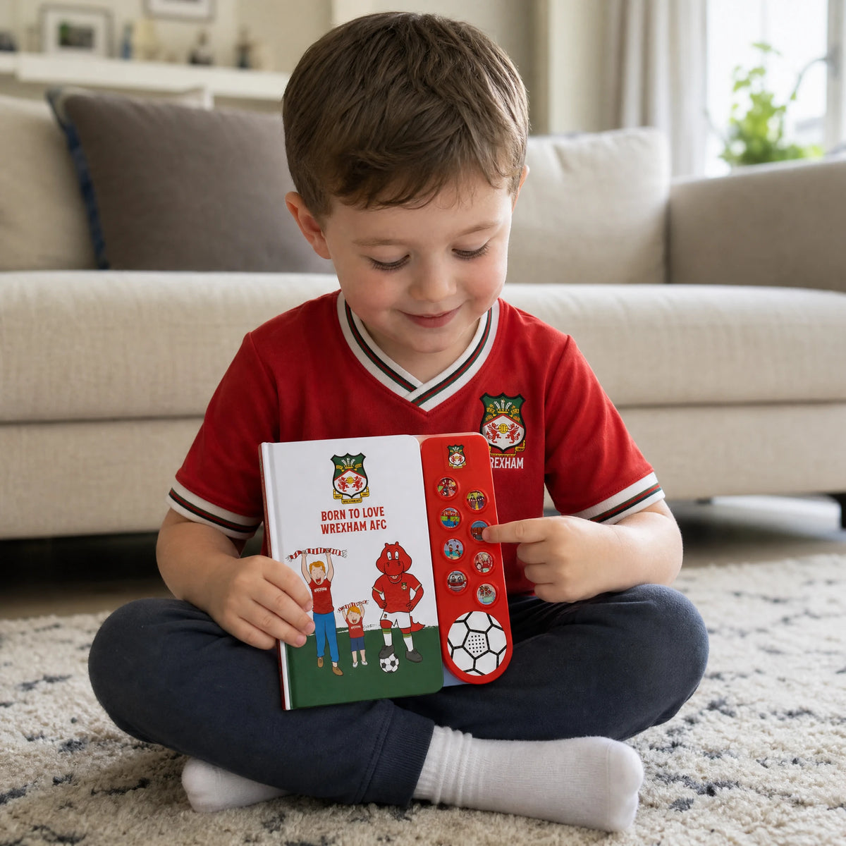 A boy in a red Wrexham AFC shirt sitting cross-legged on the rug at home, pressing one of the eight sound buttons on the Wrexham AFC Sound Book.