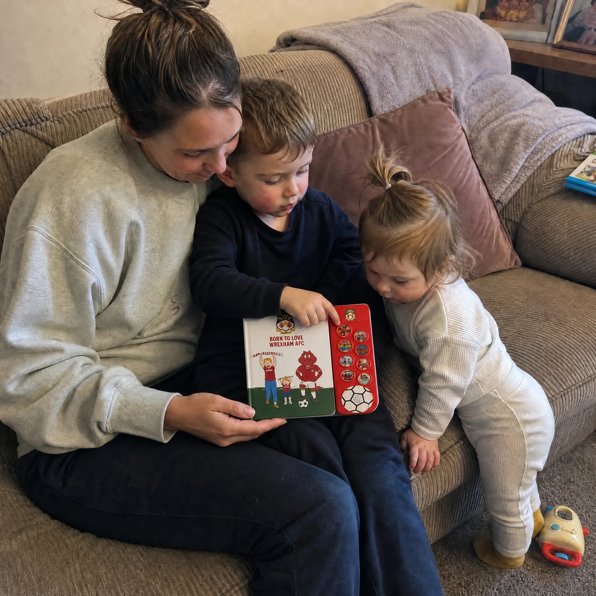 A mum on the sofa with two children, the boy pressing a sound button on the open Wrexham AFC Sound Book.